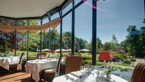 Un restaurante con mesas dispuestas para comer, amplias ventanas y vistas a un jardín.