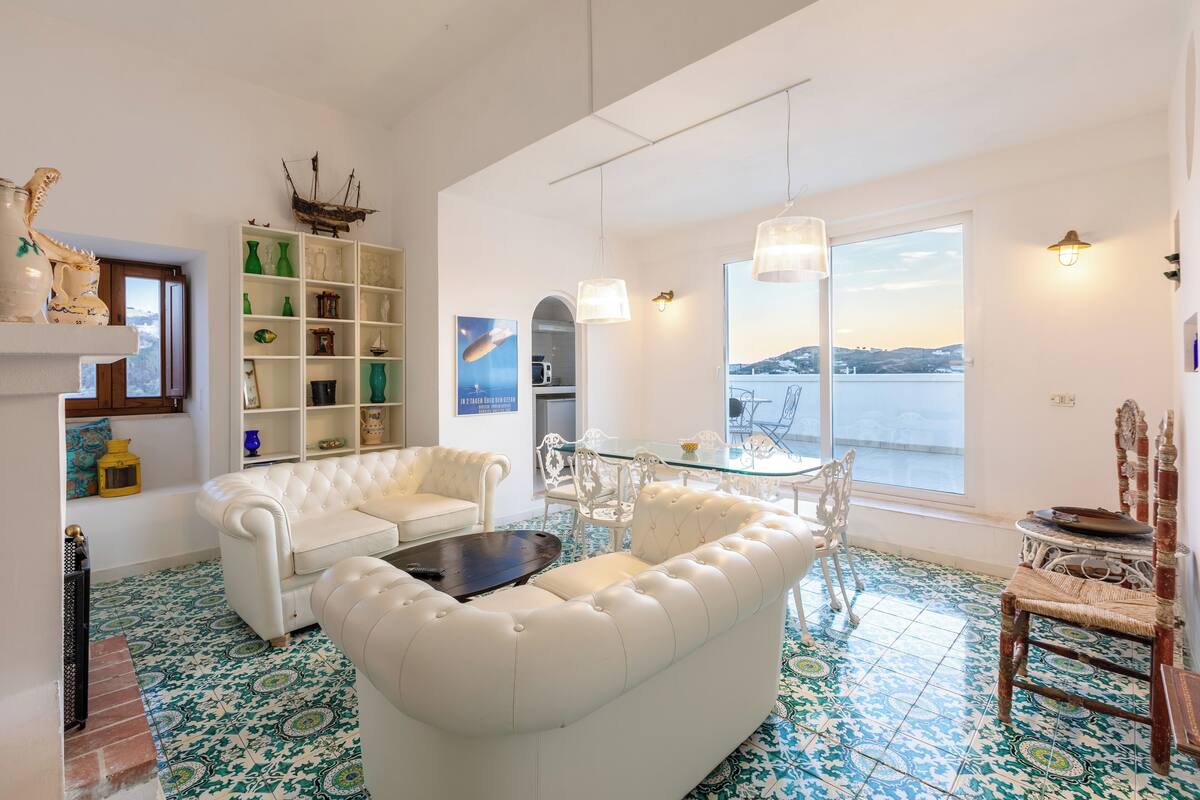 Un salon spacieux avec un canapé blanc, une table basse en verre et une vue sur la mer grâce à de grandes fenêtres.