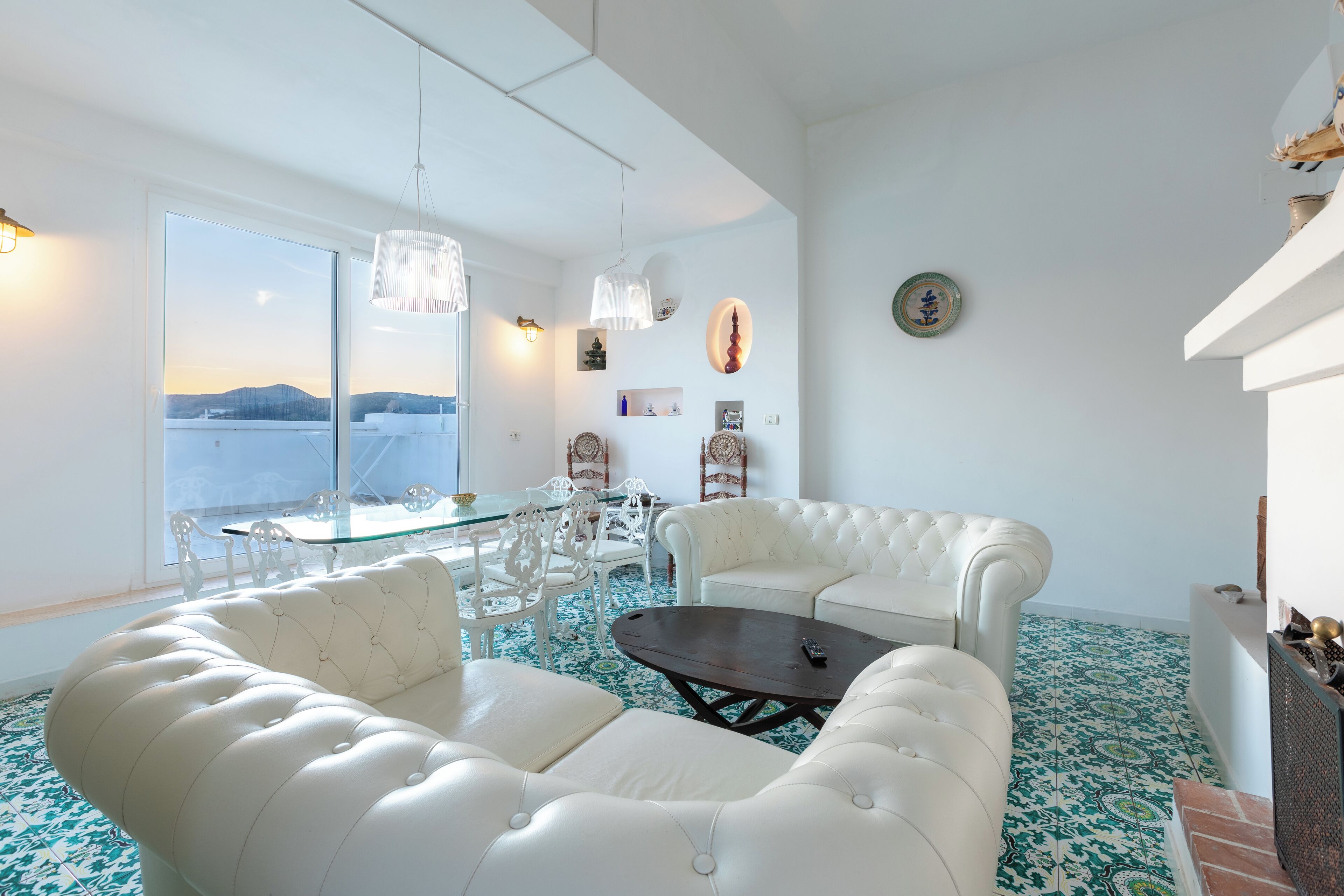 A spacious living room with a large window, white furniture, and a patterned floor.