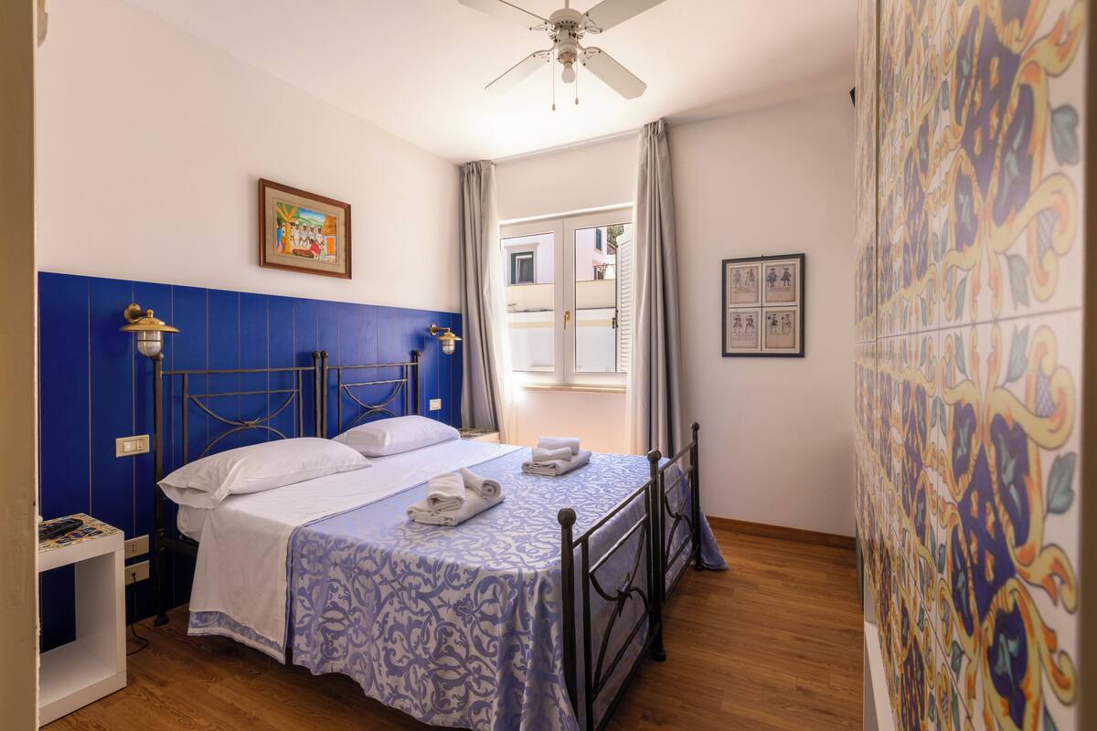 Une chambre à coucher avec un lit, une table de chevet, un ventilateur de plafond et une fenêtre avec des rideaux.
