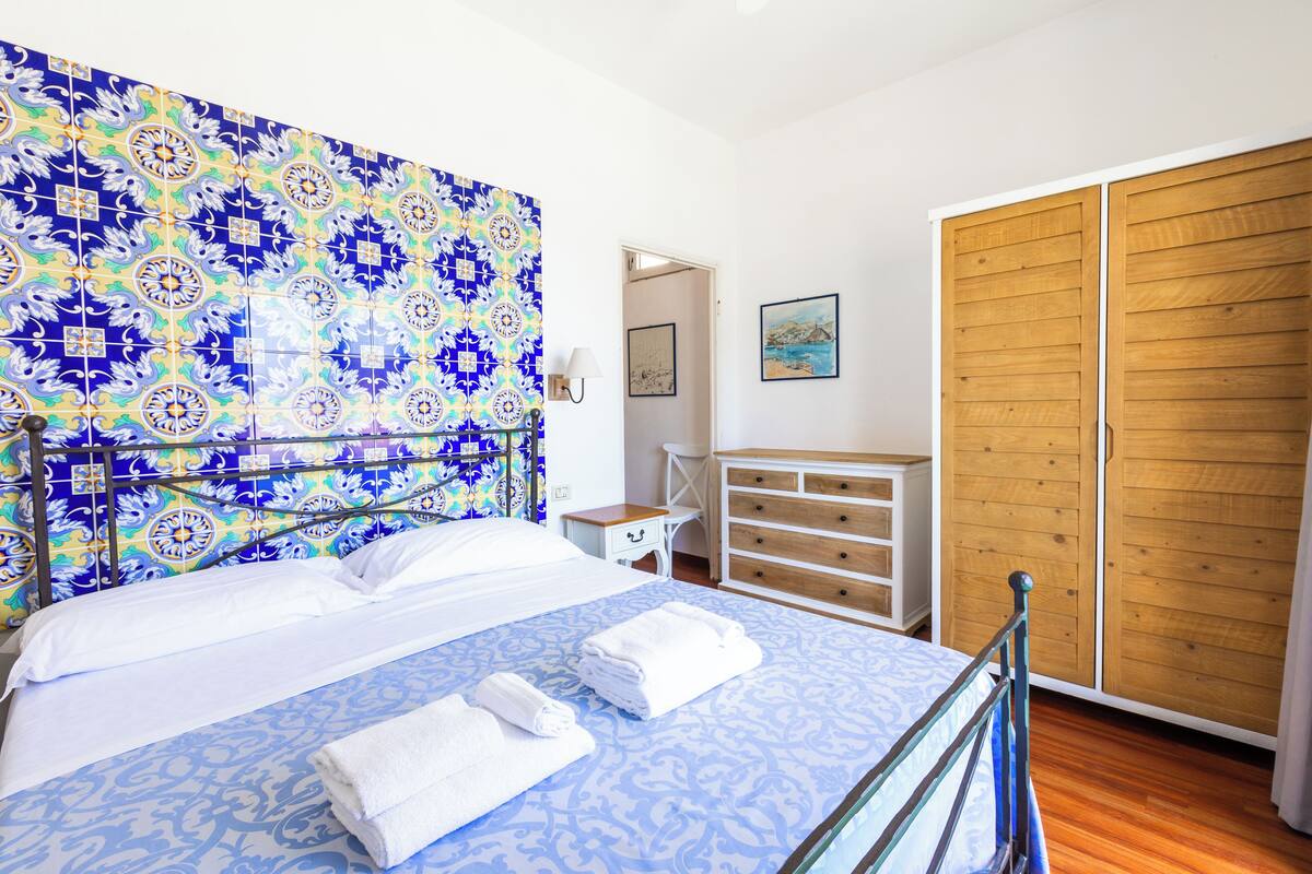 Une chambre à coucher avec une tête de lit à motifs, un lit recouvert de draps blancs, une armoire en bois et une petite table de chevet avec une lampe.