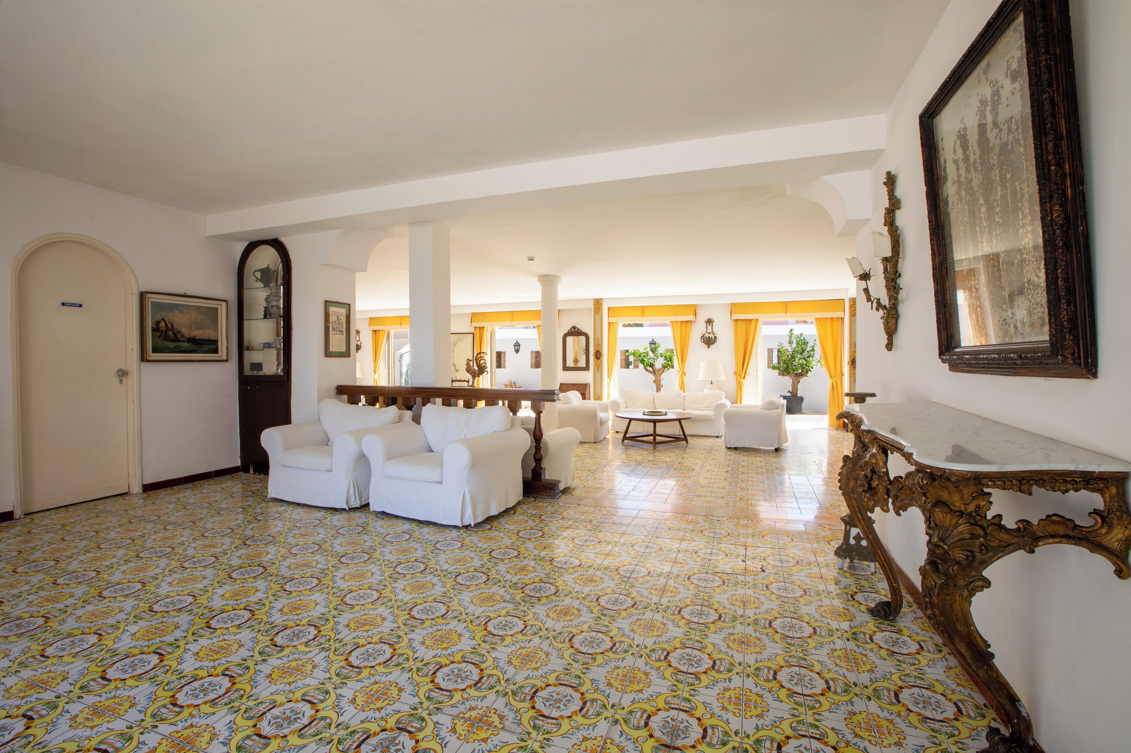 A spacious room with patterned floor tiles, white sofas, a wooden table, and a large mirror.