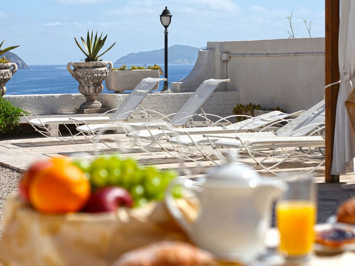 A breakfast setup with fruits, a teapot, and a glass of orange juice on a terrace with lounge chairs and a view of the sea.