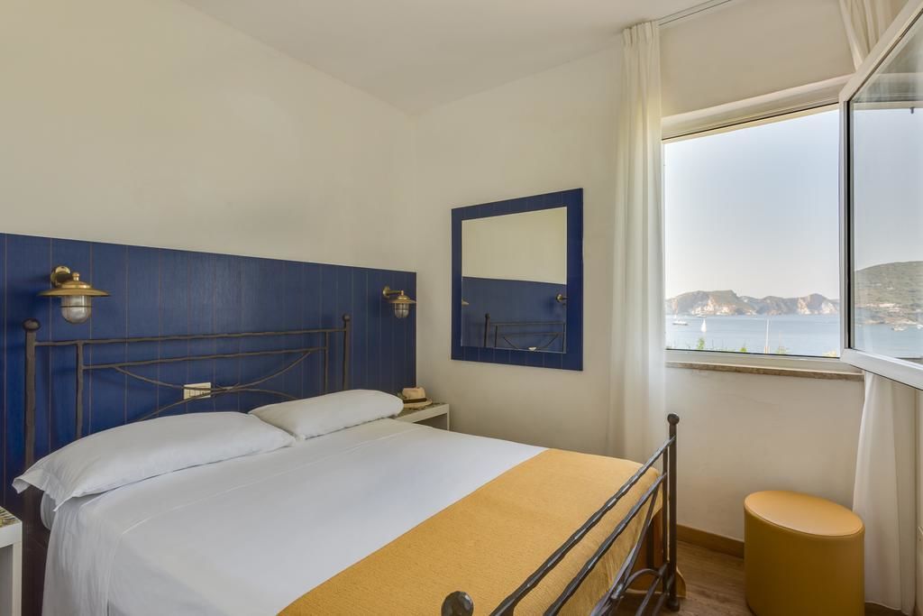 Habitación de hotel con cama, mesita de noche, espejo y ventana con vista a un cuerpo de agua y tierra distante.