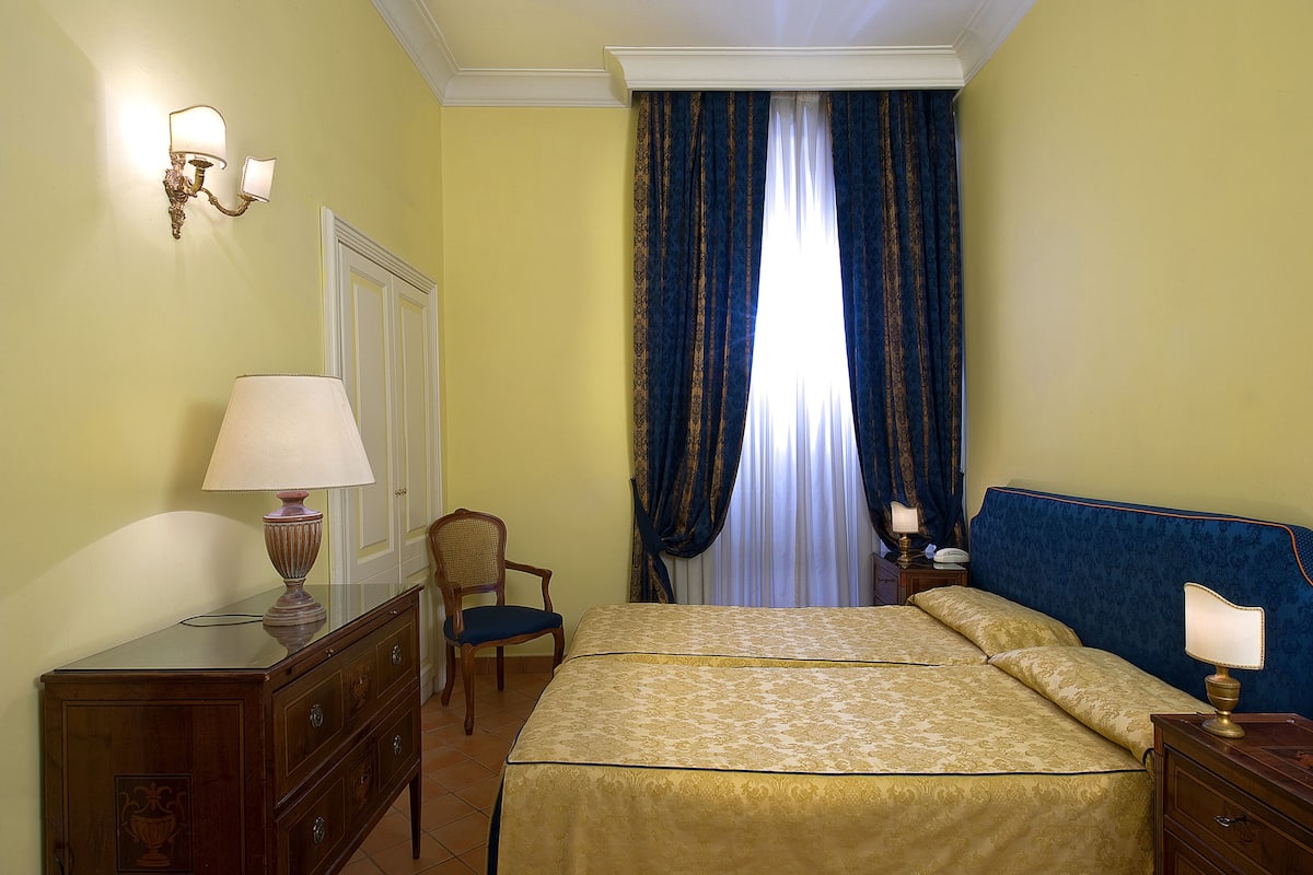 A hotel room with a bed, two bedside tables with lamps, a chair, a dresser, and a window with curtains.
