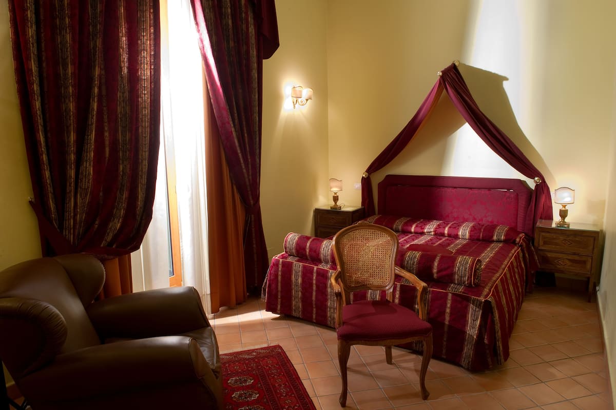 A bedroom with a canopy bed, a leather chair, a bedside table with a lamp, and a window with curtains.