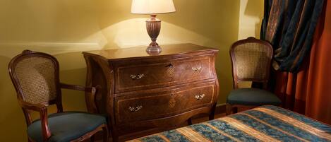 A room with a wooden dresser, two upholstered chairs, a bed with a patterned bedspread, and a lamp on the dresser.