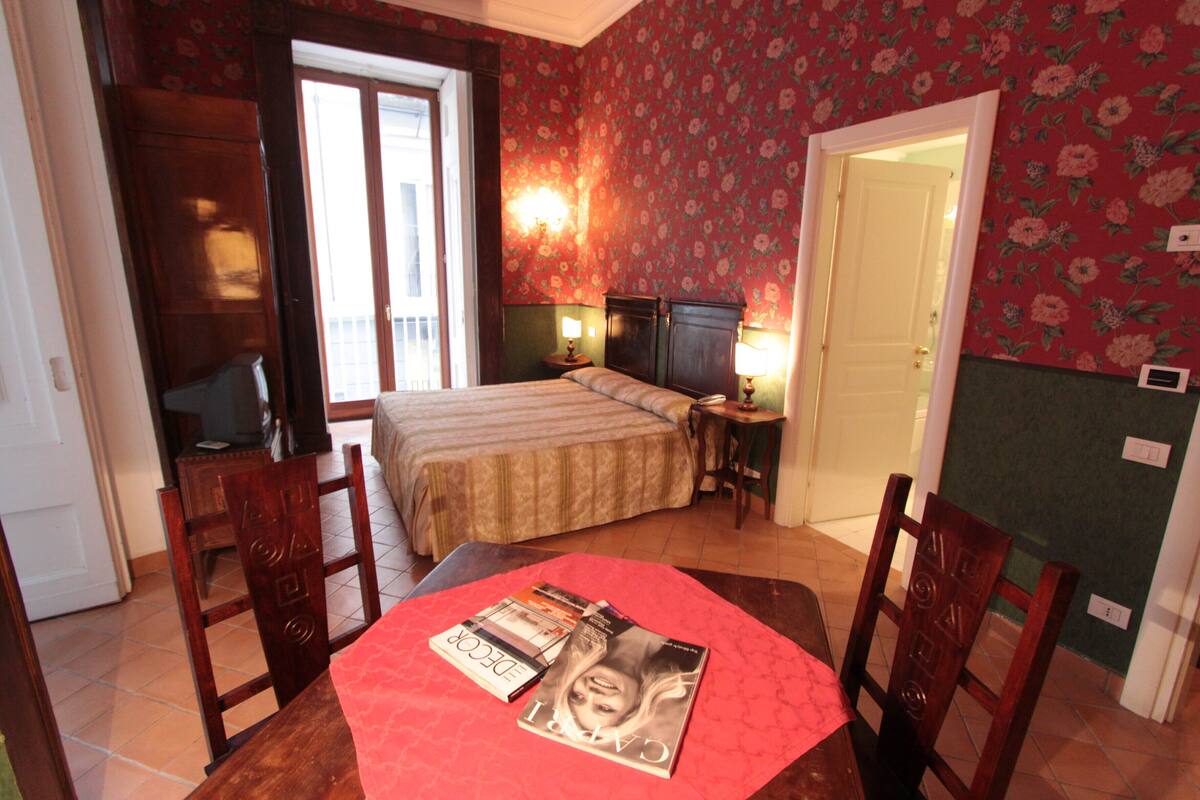 A hotel room with a bed, a desk, and a chair. There is a window with curtains, a door, and a wall with floral wallpaper.