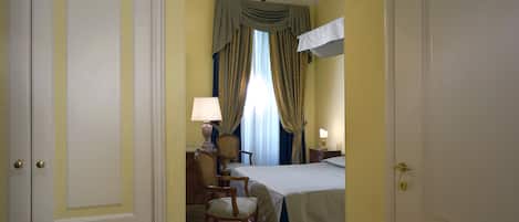 Quarto de hotel com cama, uma cadeira, escrivaninha com luminária e cortinas.
