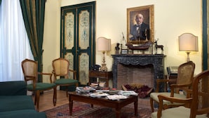 Un salón formal con chimenea, sillones tapizados en verde, una mesita de centro con revistas y un retrato sobre la chimenea.