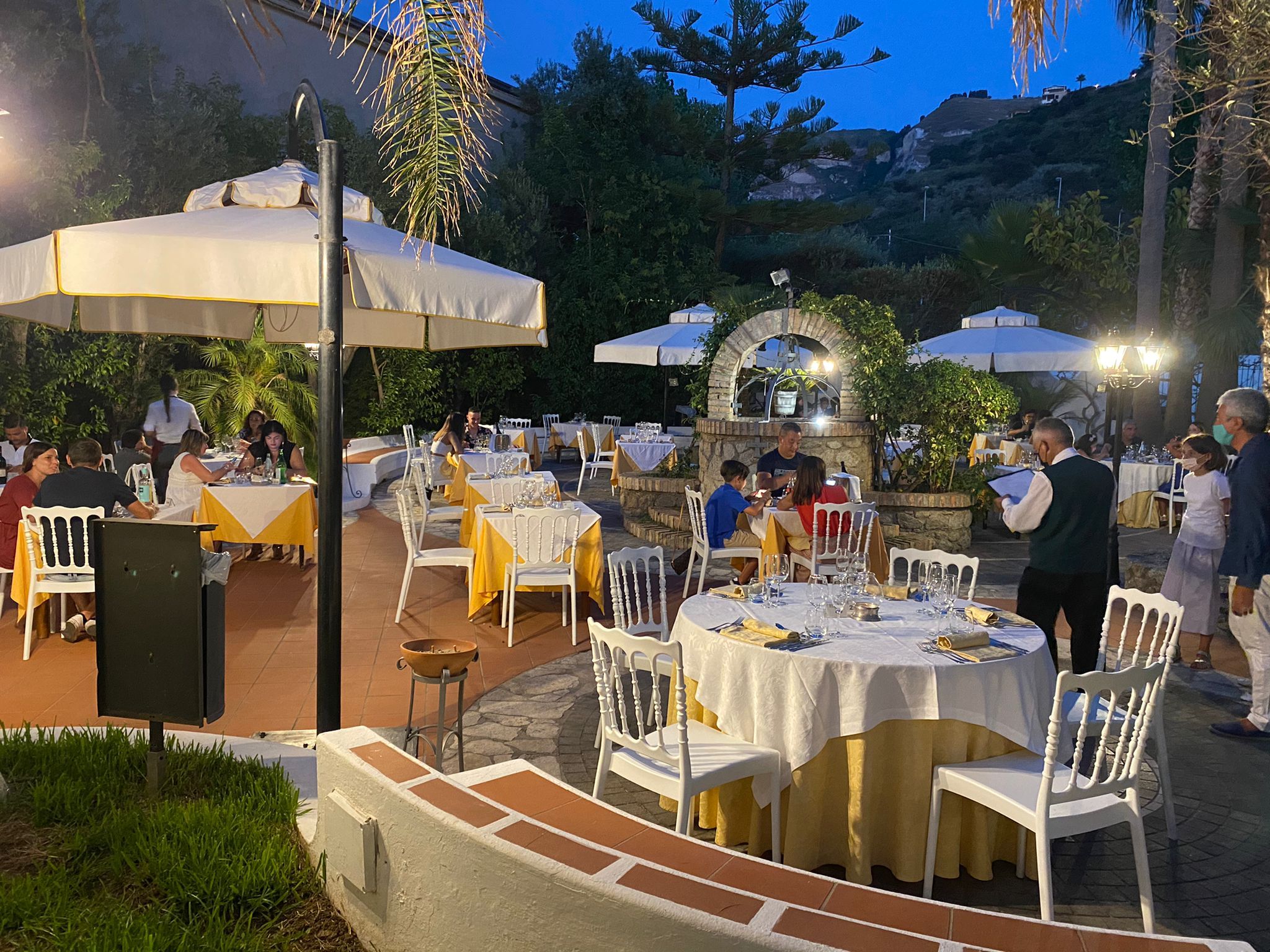 An outdoor dining area with tables and chairs, surrounded by greenery and a stone wall.