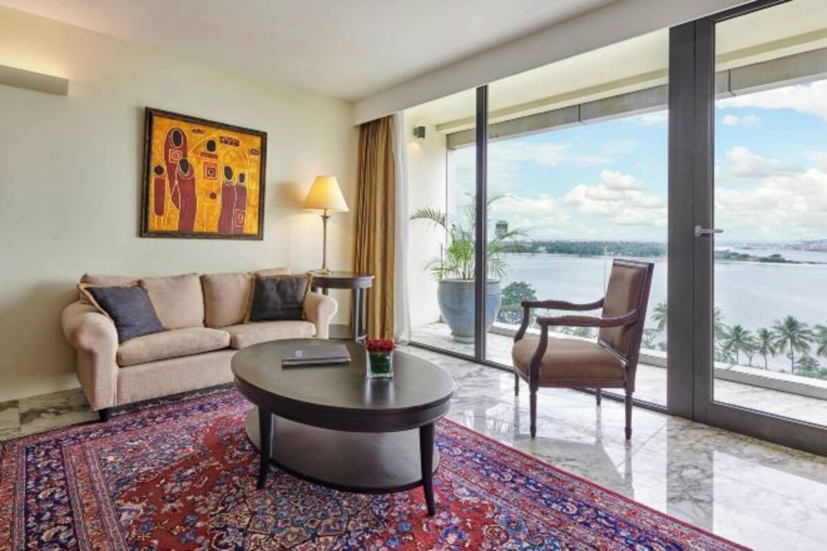 Una habitación de hotel moderna con un ventanal amplio que ofrece vistas a una masa de agua, una mesa de centro redonda, un sofá y una silla.