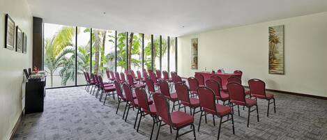 Un salón de conferencias con filas de sillas rojas, una mesa larga y ventanas amplias con vista a palmeras.