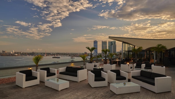 Een dakterras met witte rieten meubels, glazen tafels en uitzicht op de skyline en het water.