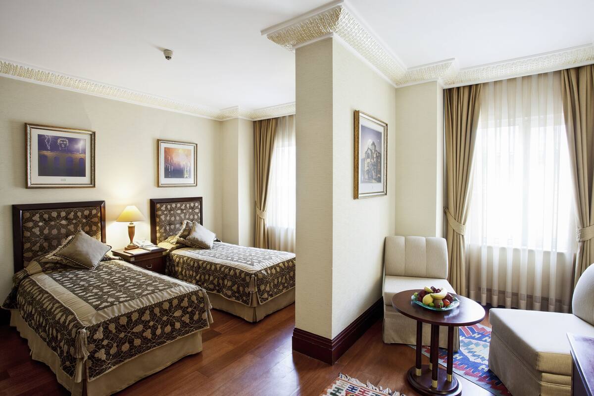 A hotel room with two beds, a sofa, a small table with fruit, and curtains.