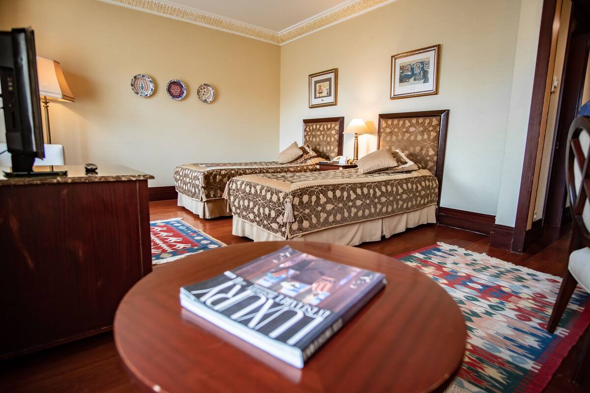 A hotel room with two beds, a desk with a lamp, a magazine on the table, and a patterned rug.