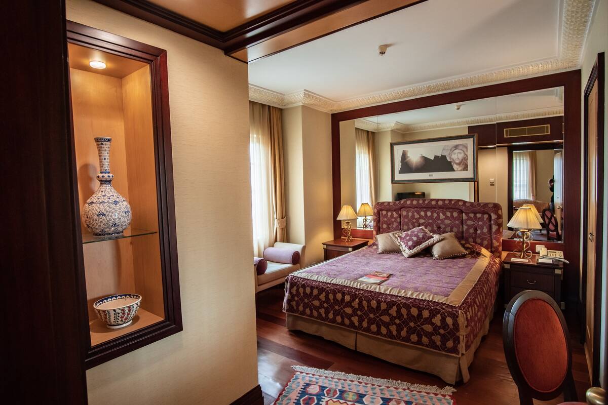 A hotel room with a large bed, a sitting area with a chair, a small table with a lamp, and a decorative vase and bowl on a shelf.