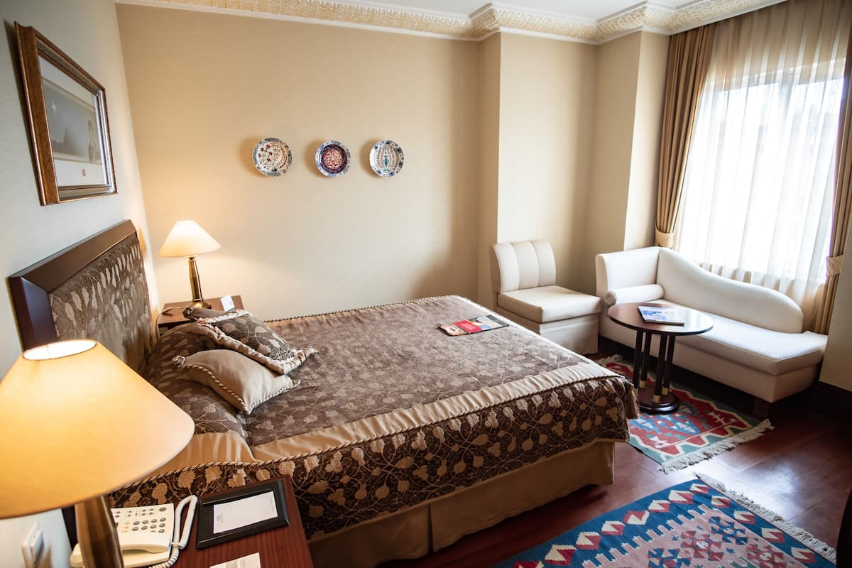 A hotel room with a large bed, a bedside table with a lamp, a chair, a small table, and a window with curtains.