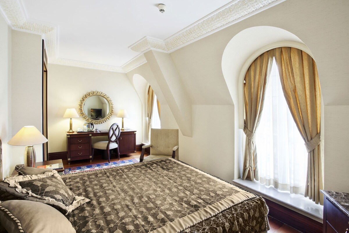 Une chambre d’hôtel avec un grand lit, un bureau avec une chaise, un miroir et une fenêtre avec des rideaux.