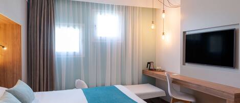 Habitación de hotel con cama, escritorio, silla, televisor y ventana con cortinas.