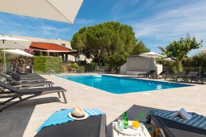 Zona de piscina con sillones de descanso, una mesa con bebidas y un árbol grande.