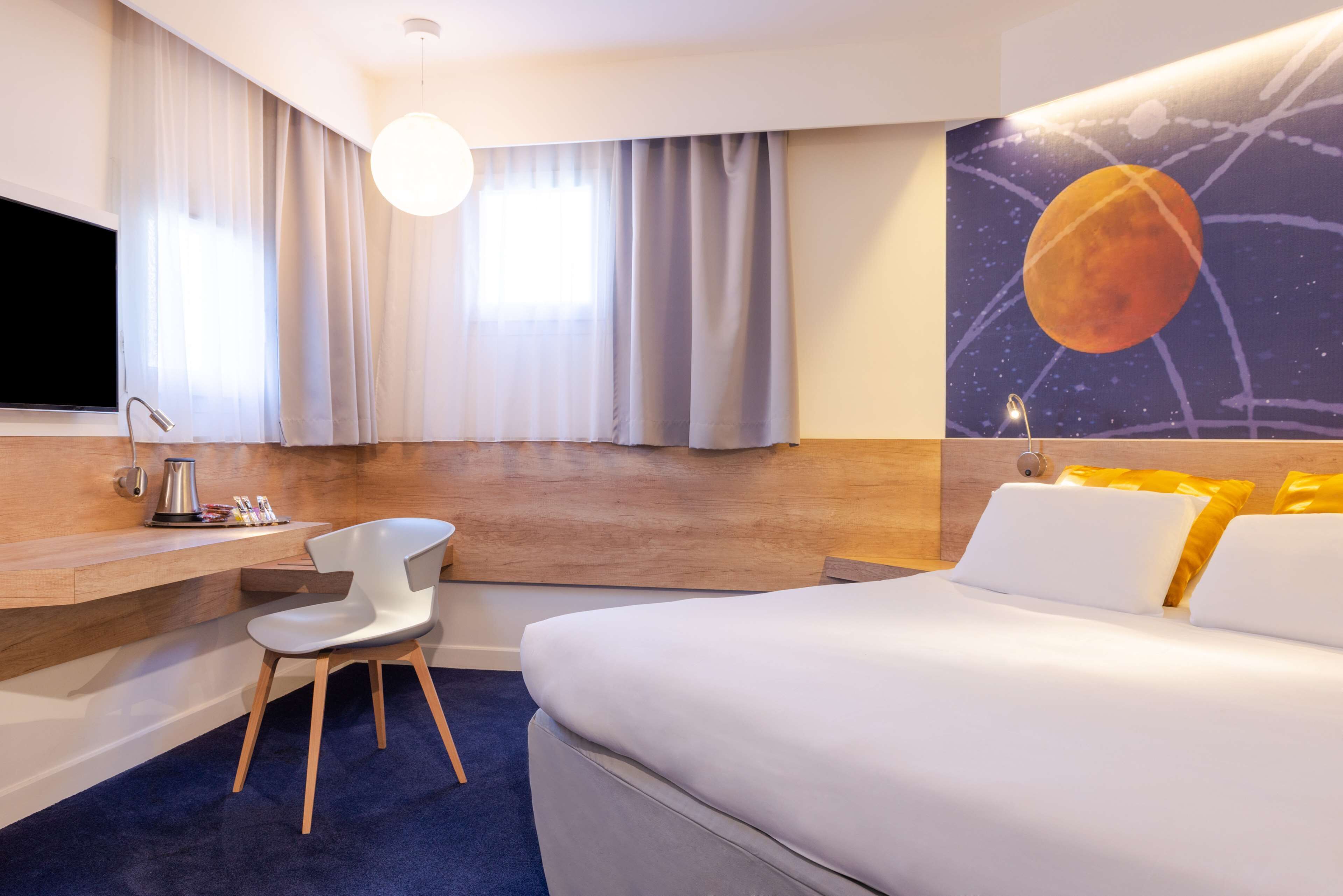 Une chambre d’hôtel avec un lit, un bureau, une chaise, une télévision et un tableau représentant une planète accroché au mur.
