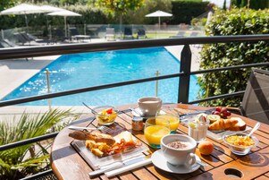Una mesa de desayuno con croissants, tostadas, fruta y bebidas, con vista a una piscina.