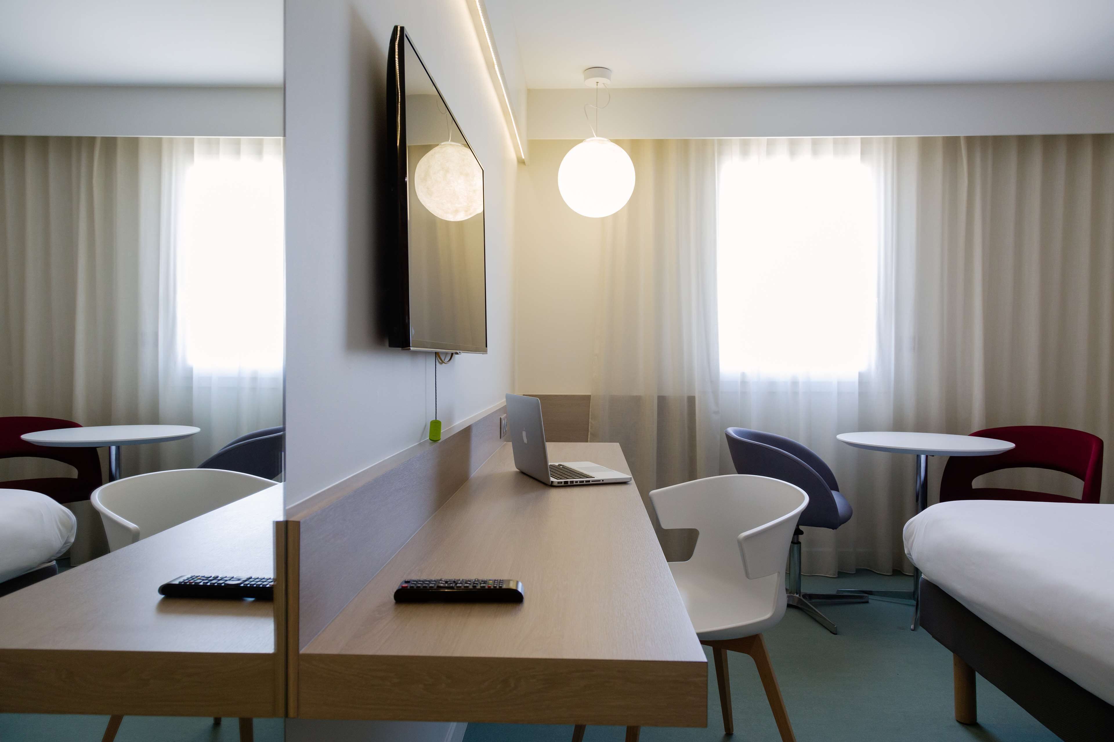 A hotel room with a bed, a desk with a laptop and remote controls, a TV mounted on the wall, and two chairs.