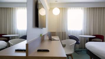 A hotel room with a bed, a desk with a laptop and remote controls, a TV mounted on the wall, and two chairs.