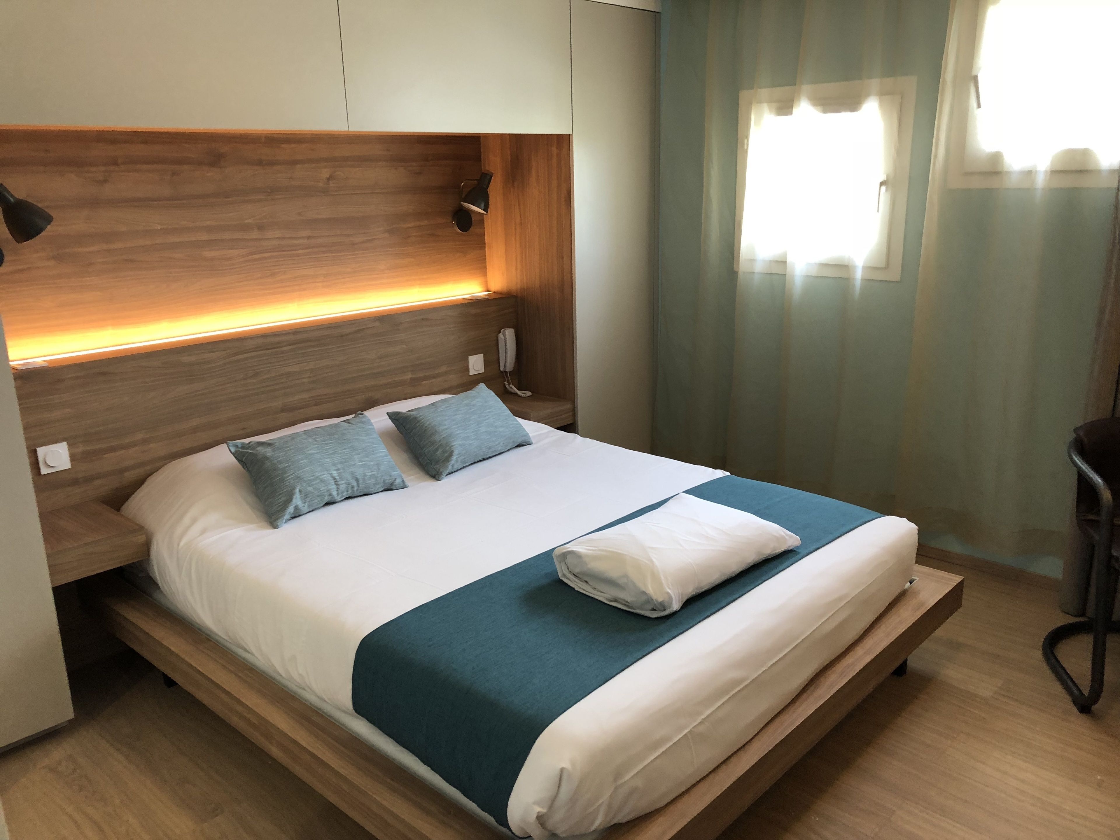 Une chambre d’hôtel moderne avec une tête de lit en bois, un téléphone sur la table de chevet et une fenêtre avec des rideaux.