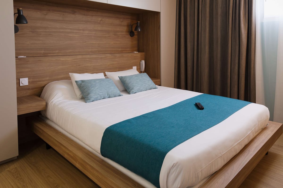 Ein ordentlich bezogenes Bett mit einer türkisblauen Tagesdecke, einem Holz-Kopfteil und Nachttischlampen.