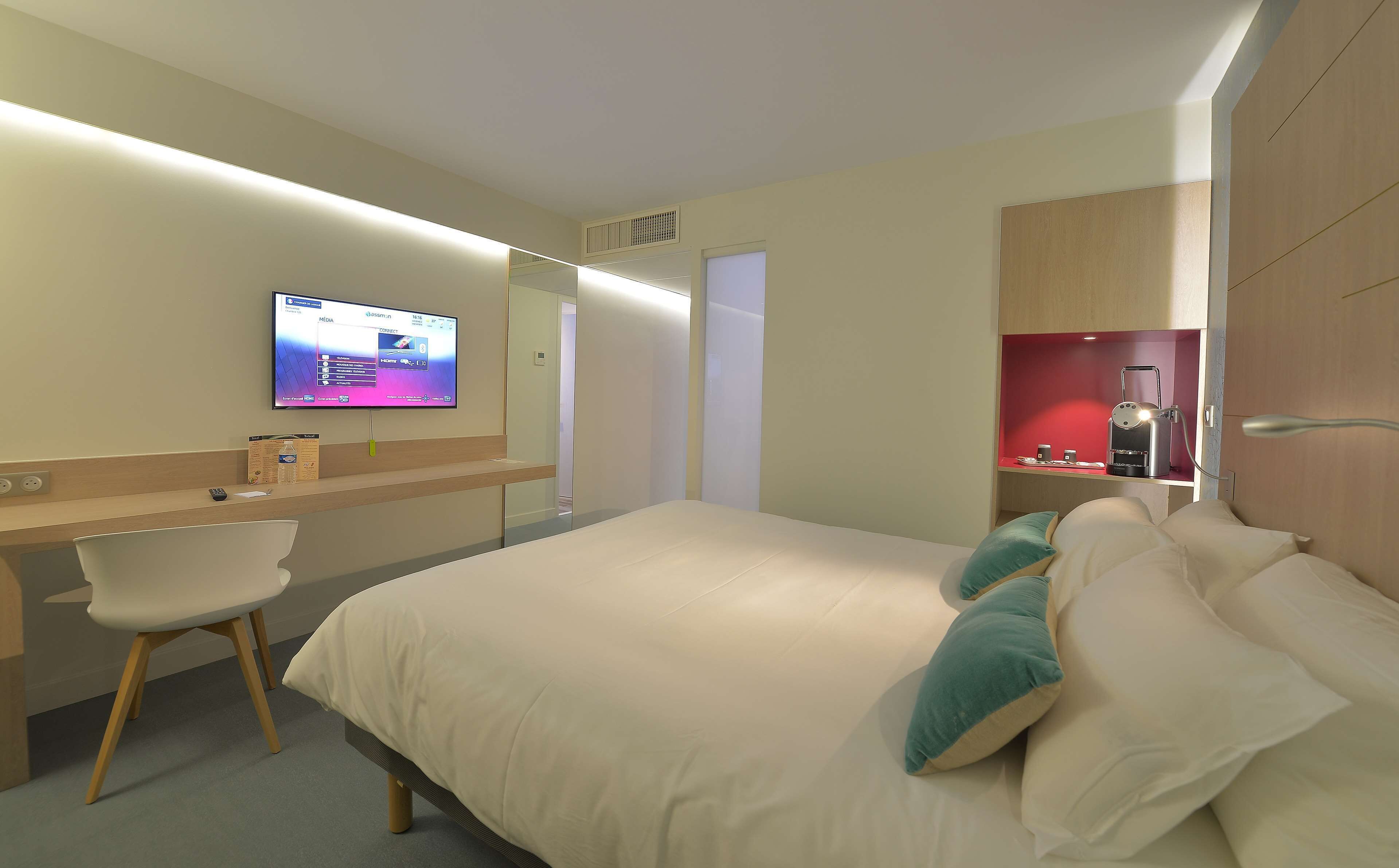 A hotel room with a bed, a desk, a chair, a television, and a red shelf with toiletries.