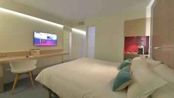 Una habitación de hotel con cama, escritorio, silla, televisor y estante rojo con artículos de aseo.
