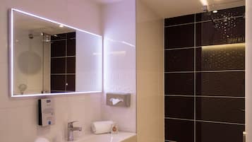 A modern bathroom with a large mirror, a sink, a towel dispenser, and a shower area.