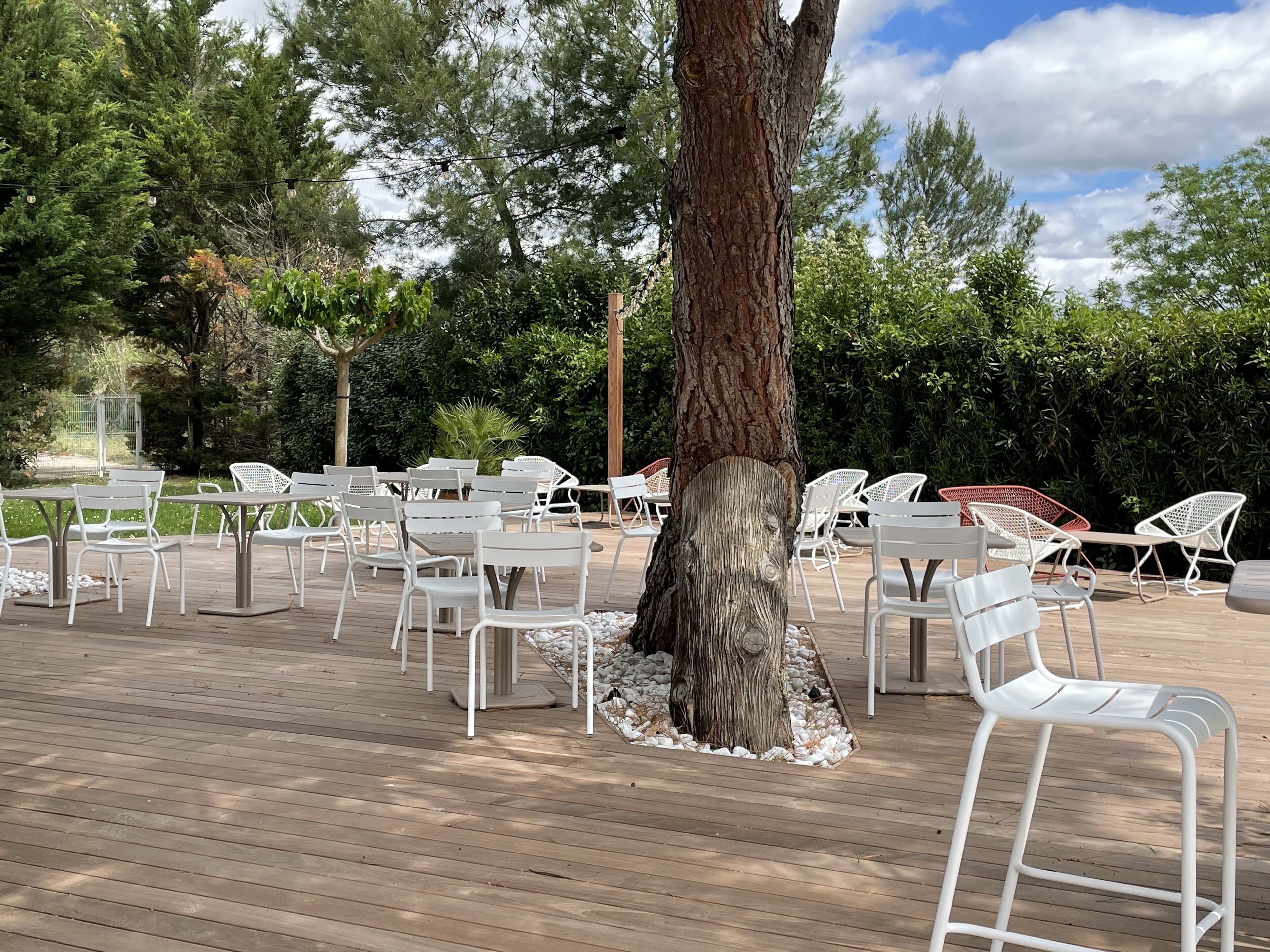 Un patio al aire libre con sillas y mesas de metal blanco, rodeado de árboles y follaje.