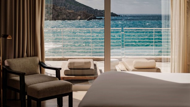 Habitación de hotel con balcón que da al mar, una cama, una silla y una pequeña mesa.