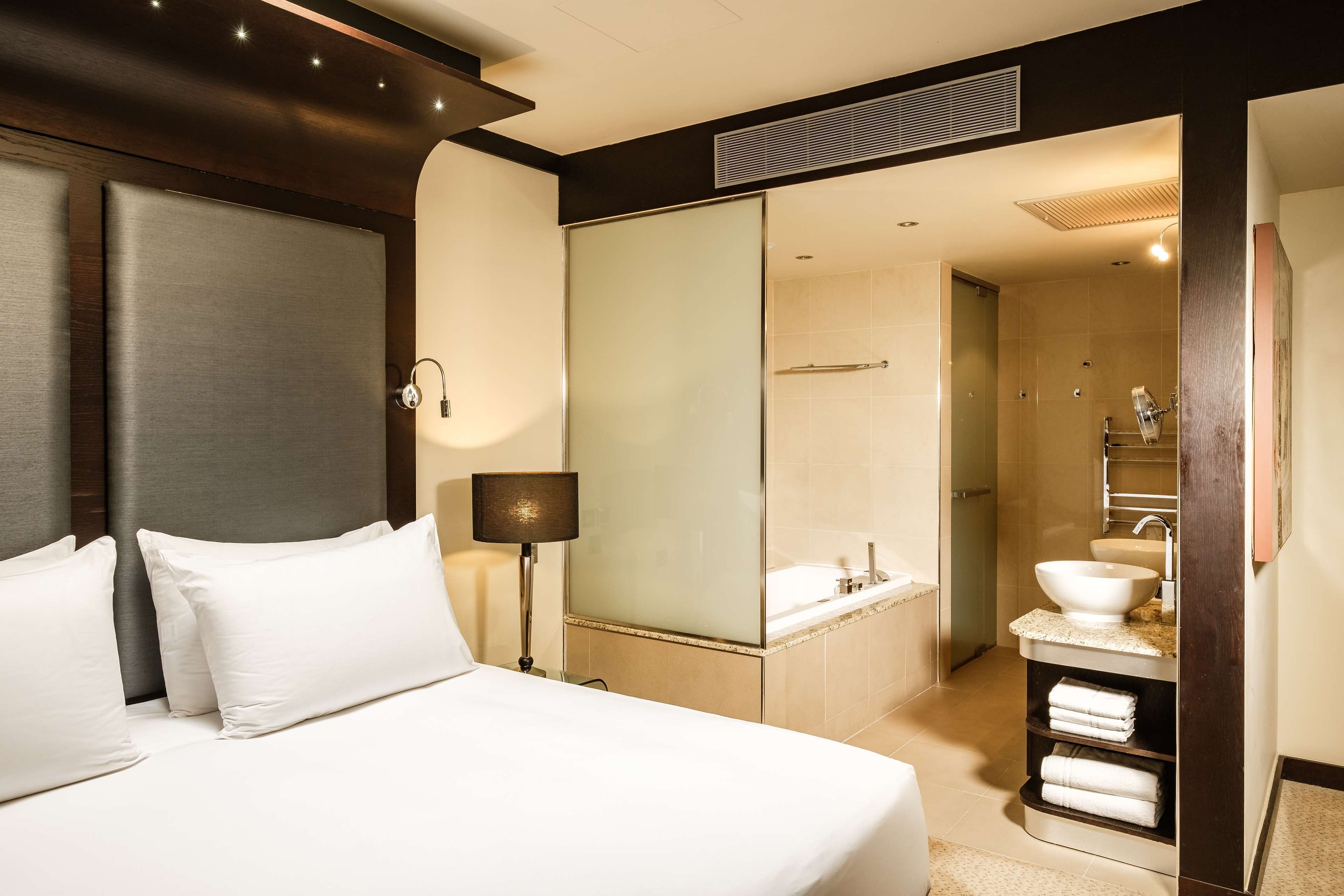 A hotel room with a bed, a bedside lamp, a large mirror, and a bathroom with a sink and towel rack.