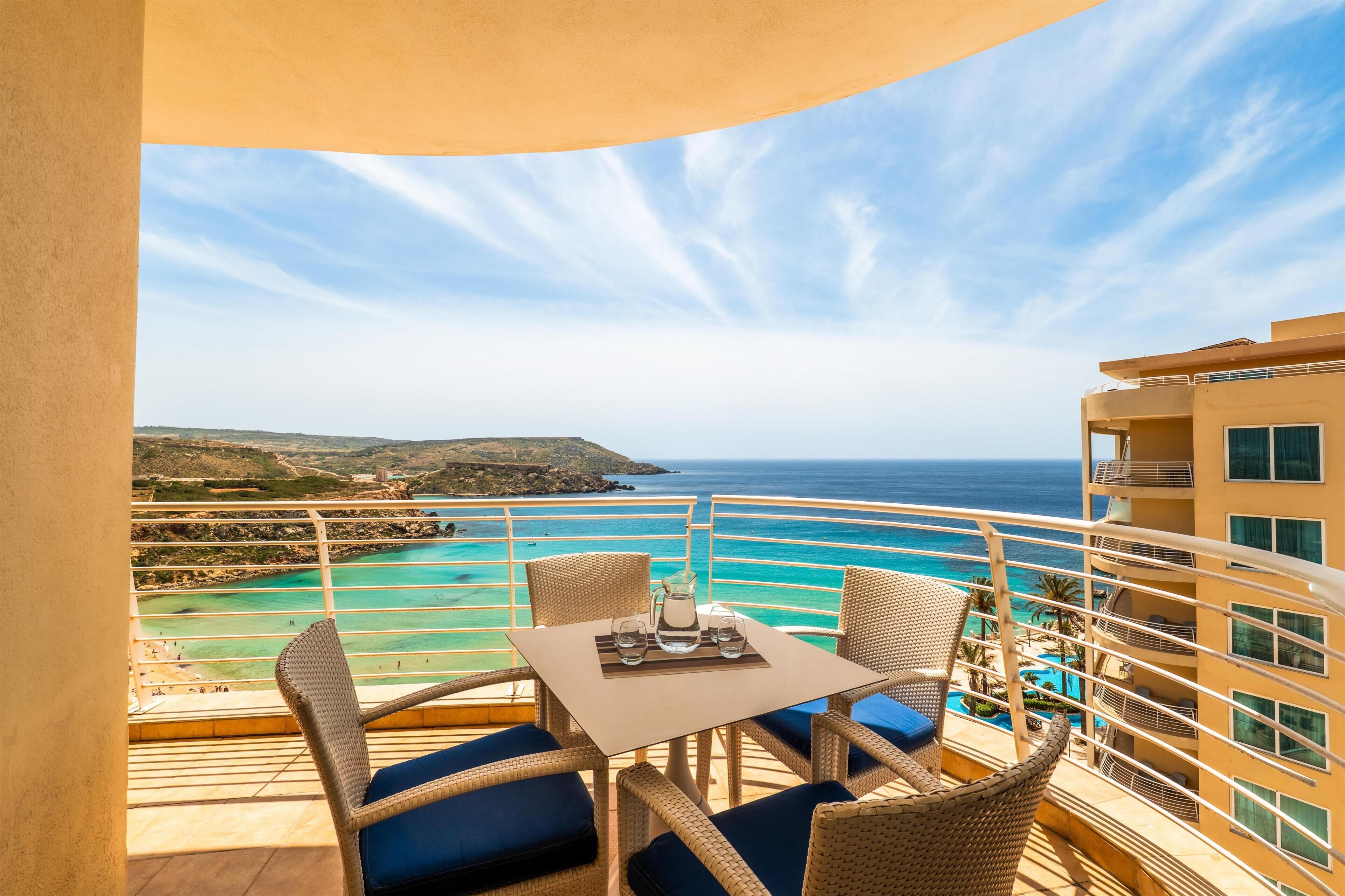 Un balcon agrémenté de meubles en osier, une table avec une carafe et des verres, et une vue sur la mer et la côte.