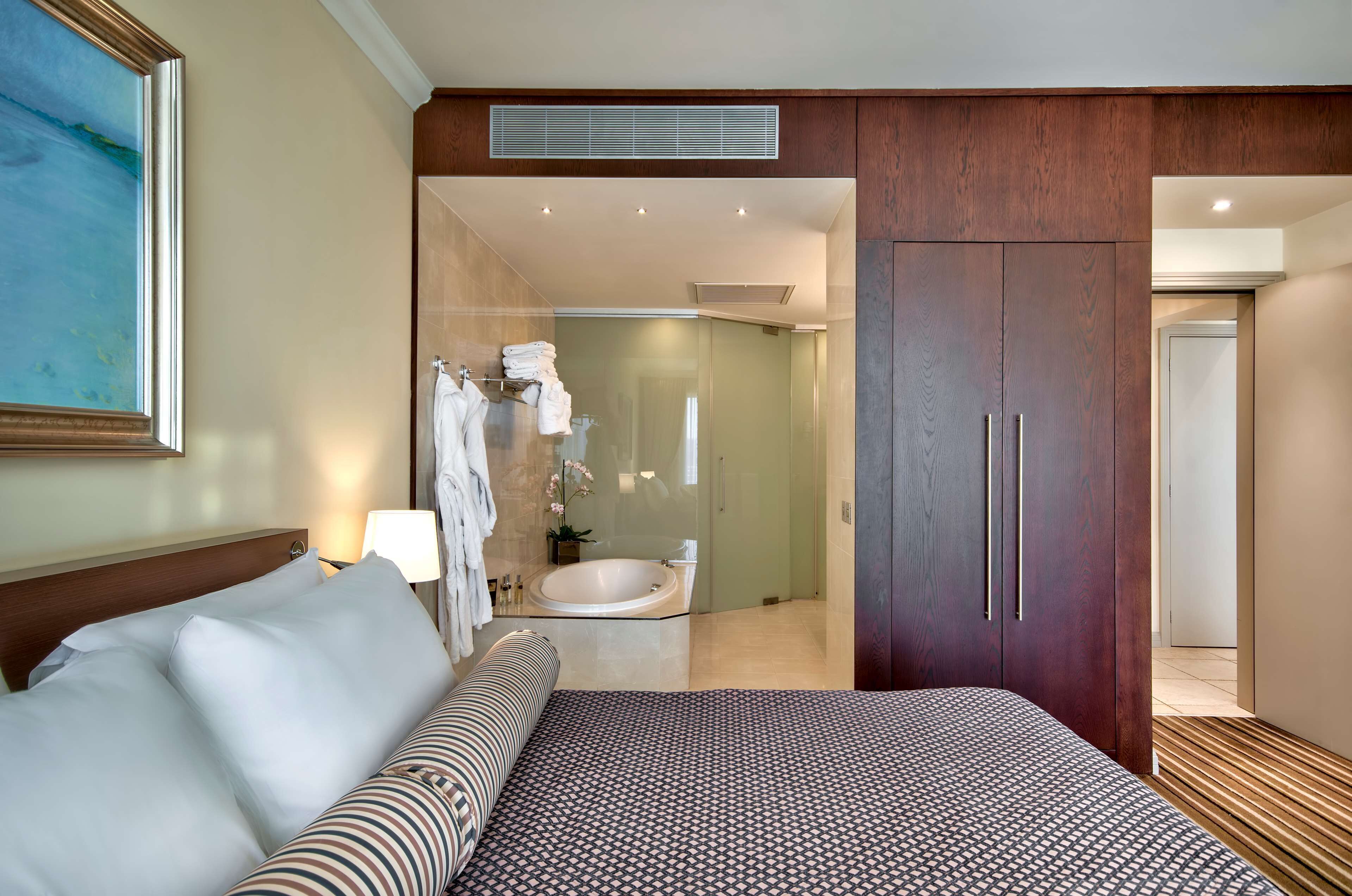 A hotel room with a large bed, a bedside table with a lamp, a shower area, and a wardrobe.