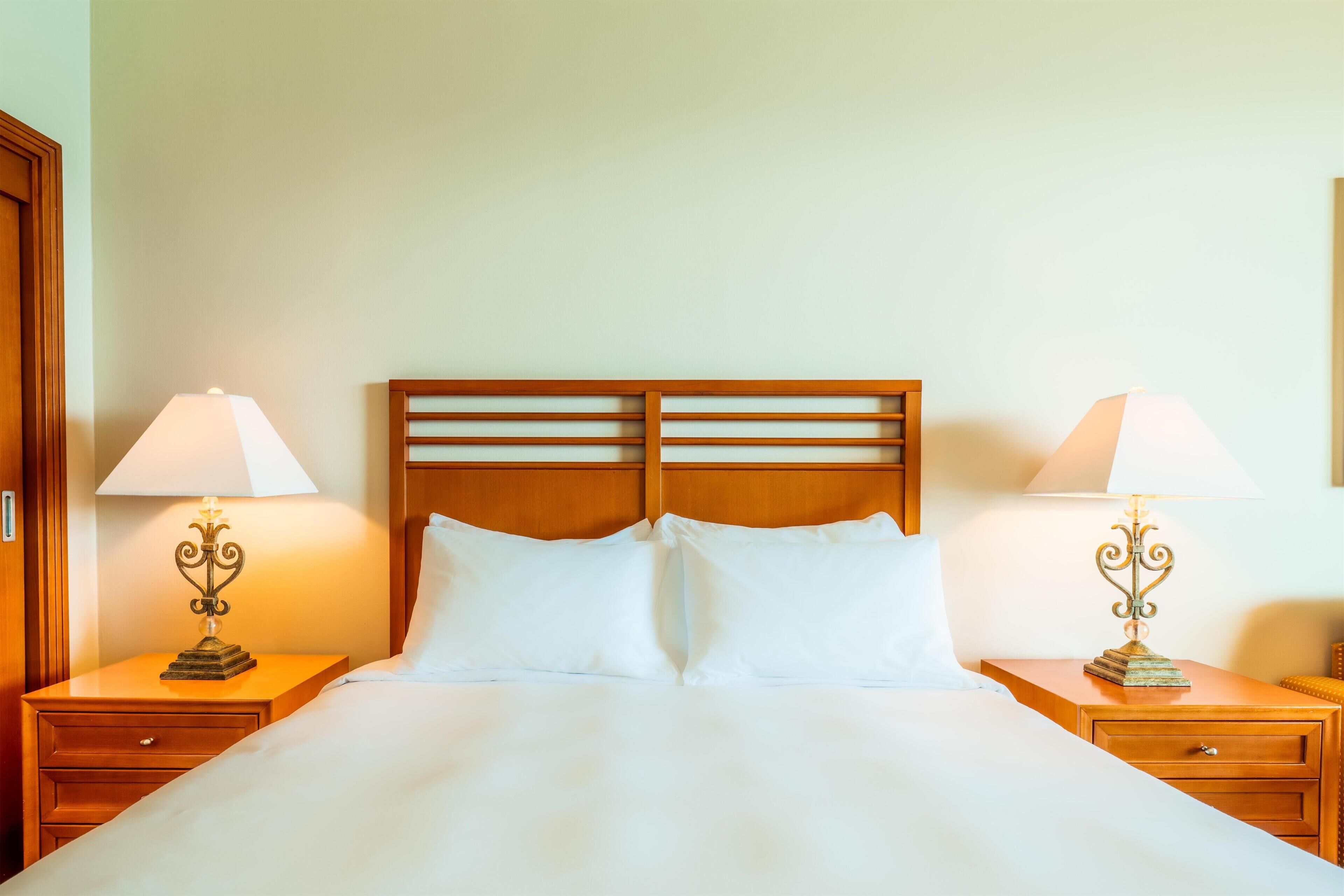 A hotel room with a wooden headboard, two bedside tables with lamps, a bed with white linens, and a wooden nightstand.