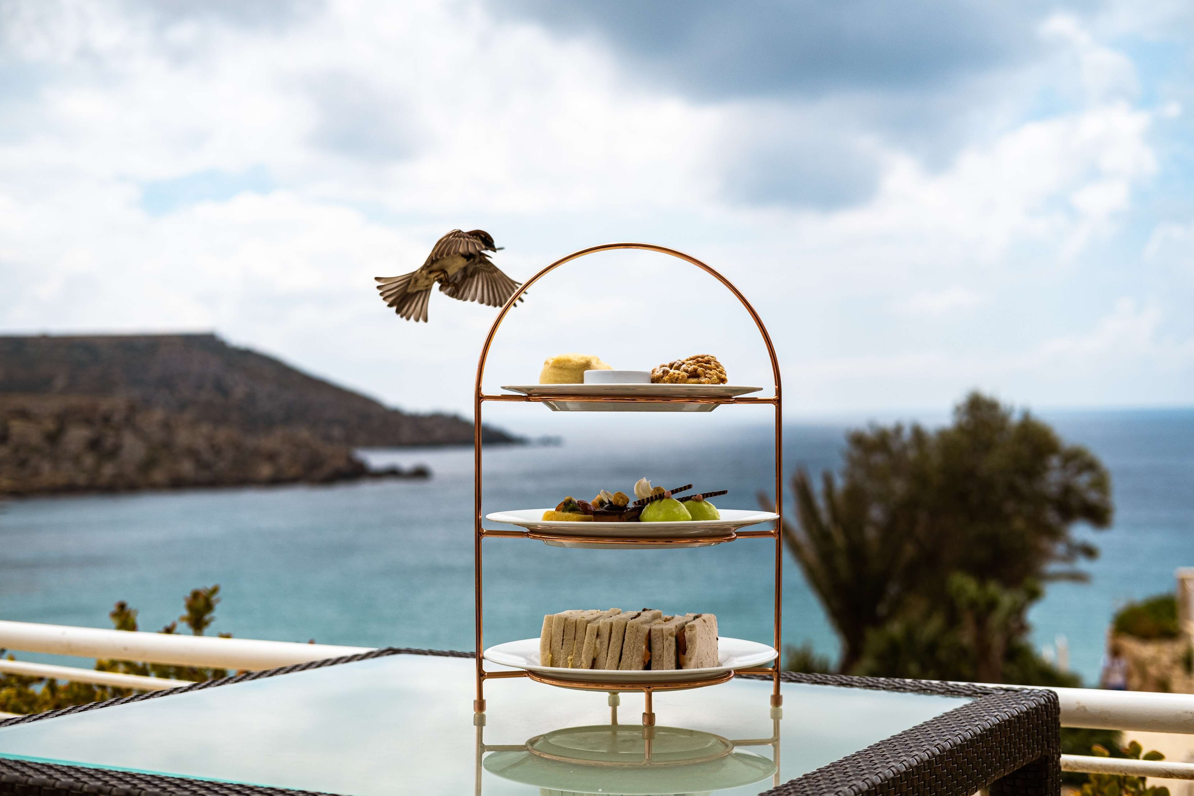 A tiered dessert stand with various treats on a glass table, overlooking a scenic coastal view.