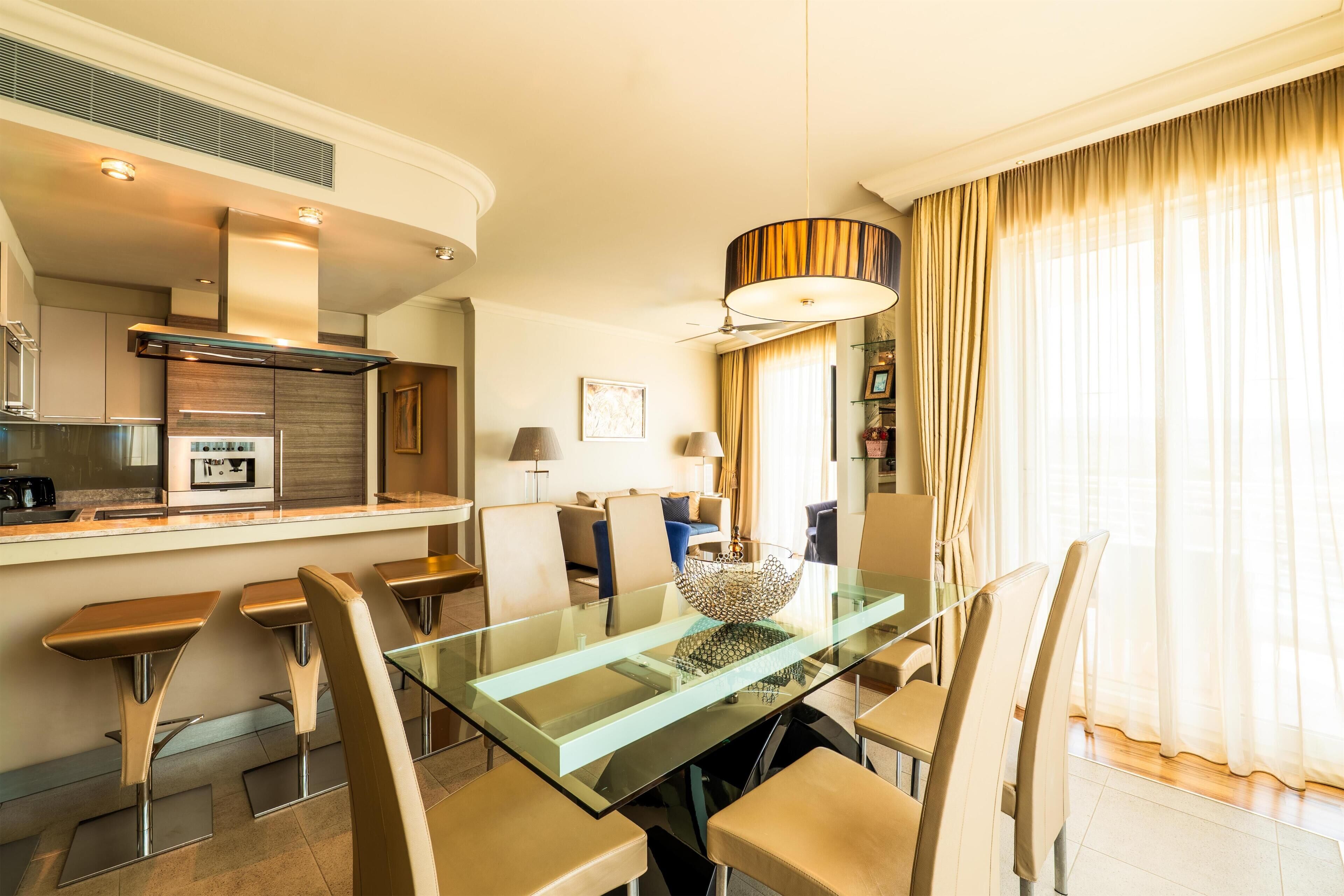 A modern kitchen with a glass dining table, bar stools, and a pendant light.