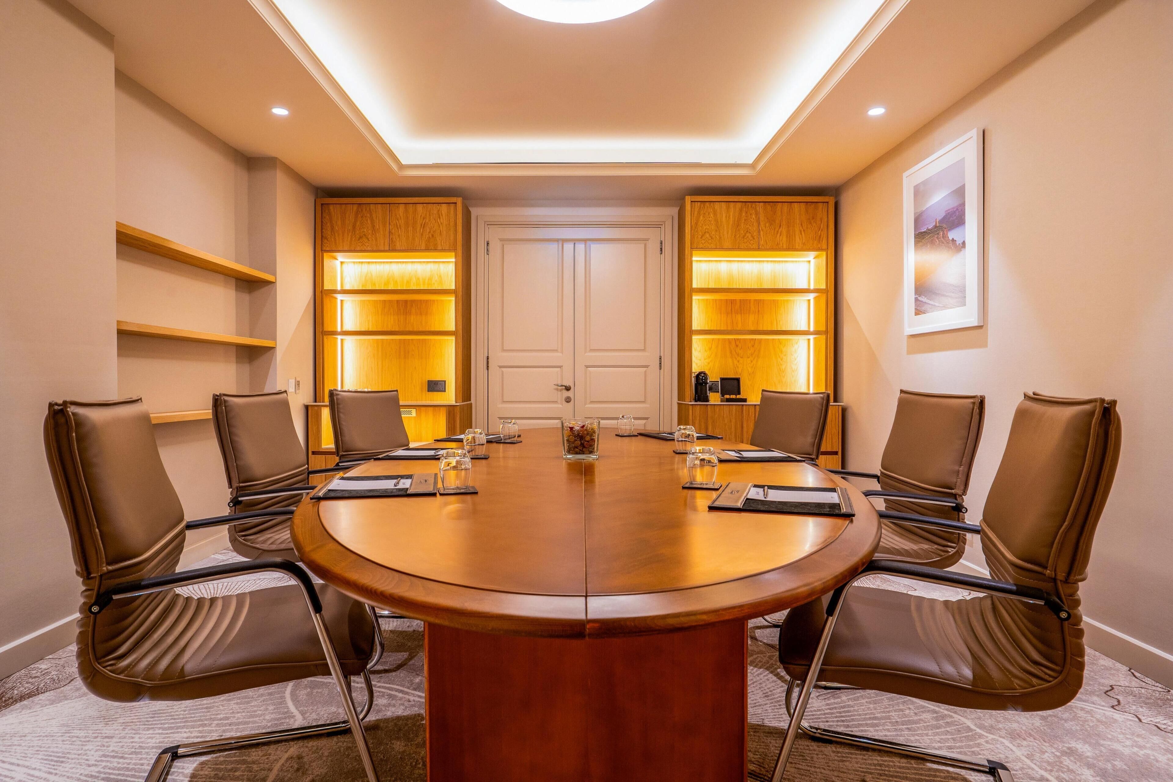 Une salle de conférence dotée d’une table ronde, de fauteuils en cuir marron, d’étagères en bois avec des panneaux rétroéclairés et d’un tableau encadré au mur.