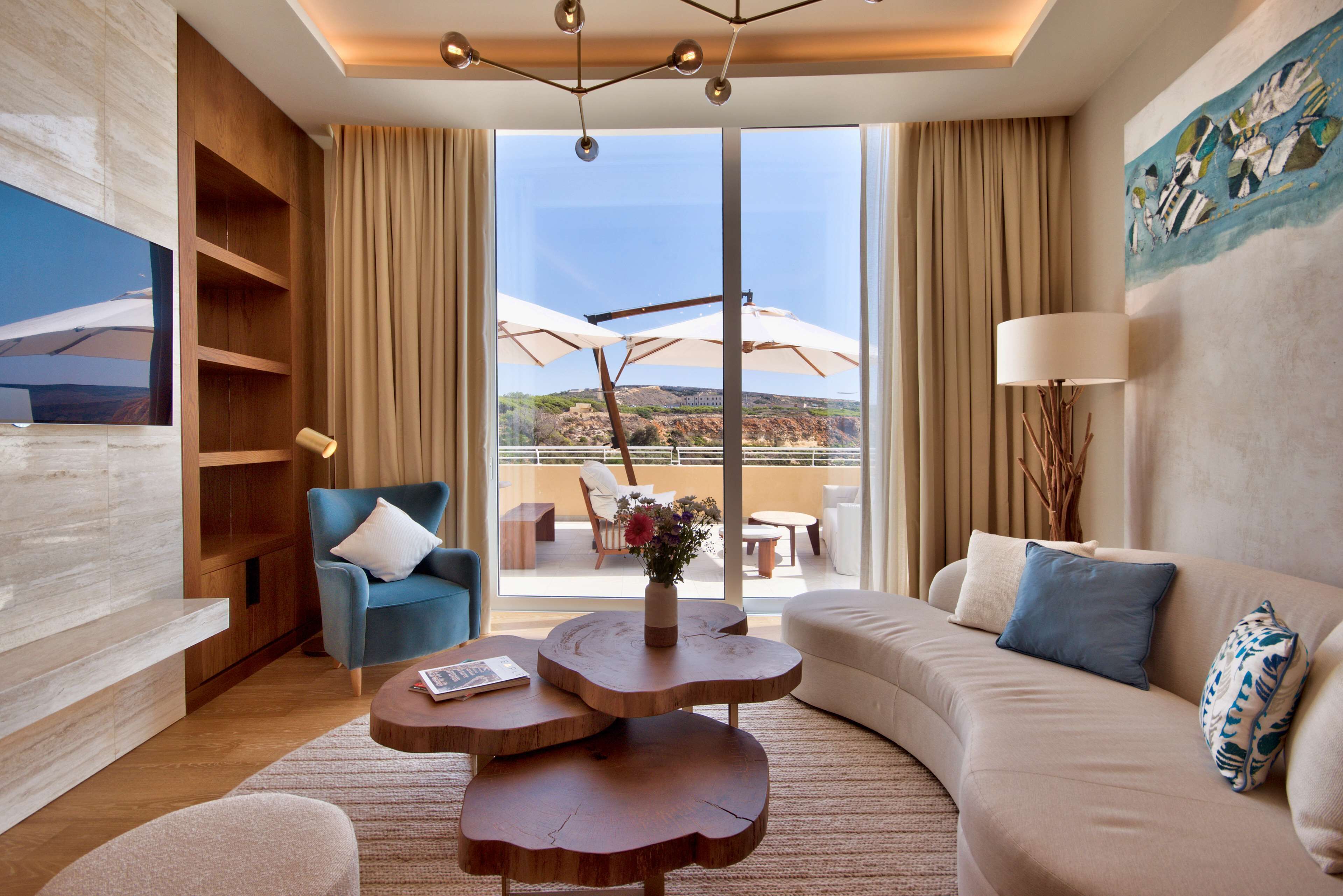 Una sala de estar moderna con un sofá, un sillón azul, una mesa de centro de madera y vistas al exterior a través de grandes ventanales.