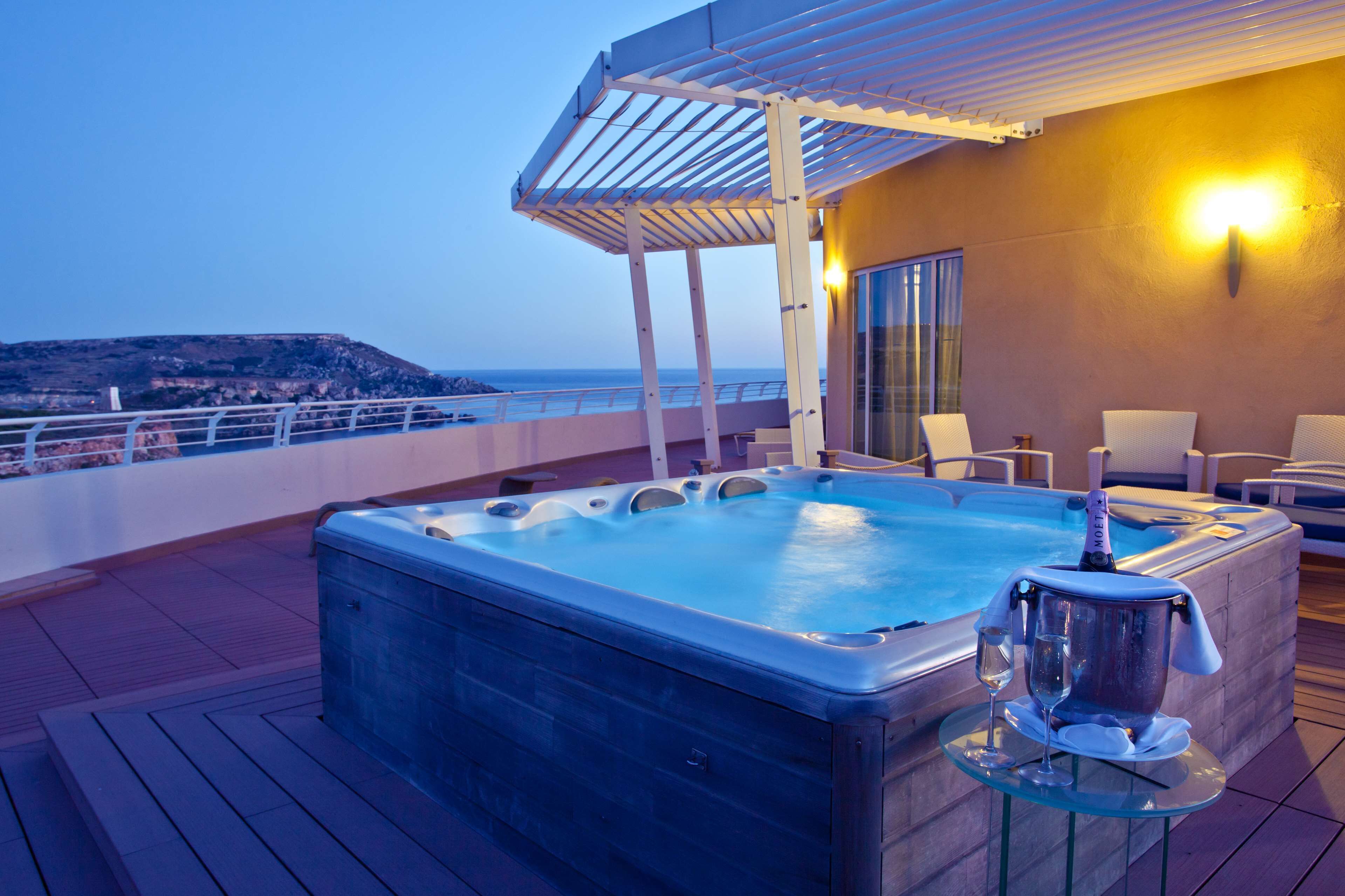 A hot tub on a deck with a view of the sea, a covered seating area, and a table with a bottle and glass.