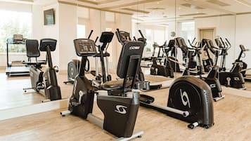 A gym with various exercise machines, including stationary bikes and treadmills, arranged in a spacious room with large windows and a wooden floor.