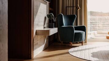 Un moderno salón con un sillón azul oscuro, suelo de madera, pared decorativa de mármol, una planta pequeña y vistas al exterior a través de amplias ventanas.