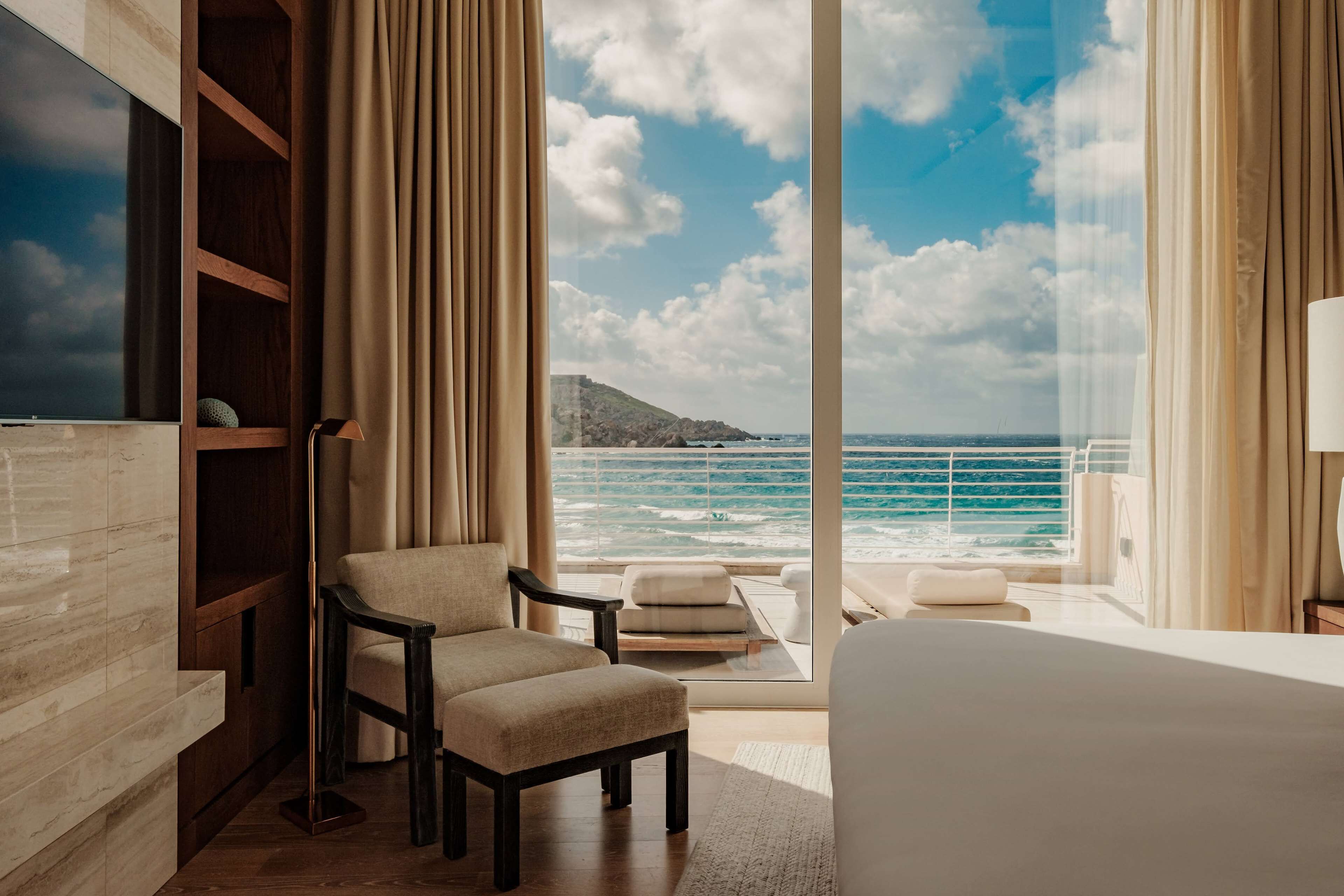 Habitación de hotel con una cama grande, un área de estar con vista al océano, una estantería de madera y un amplio ventanal con cortinas.