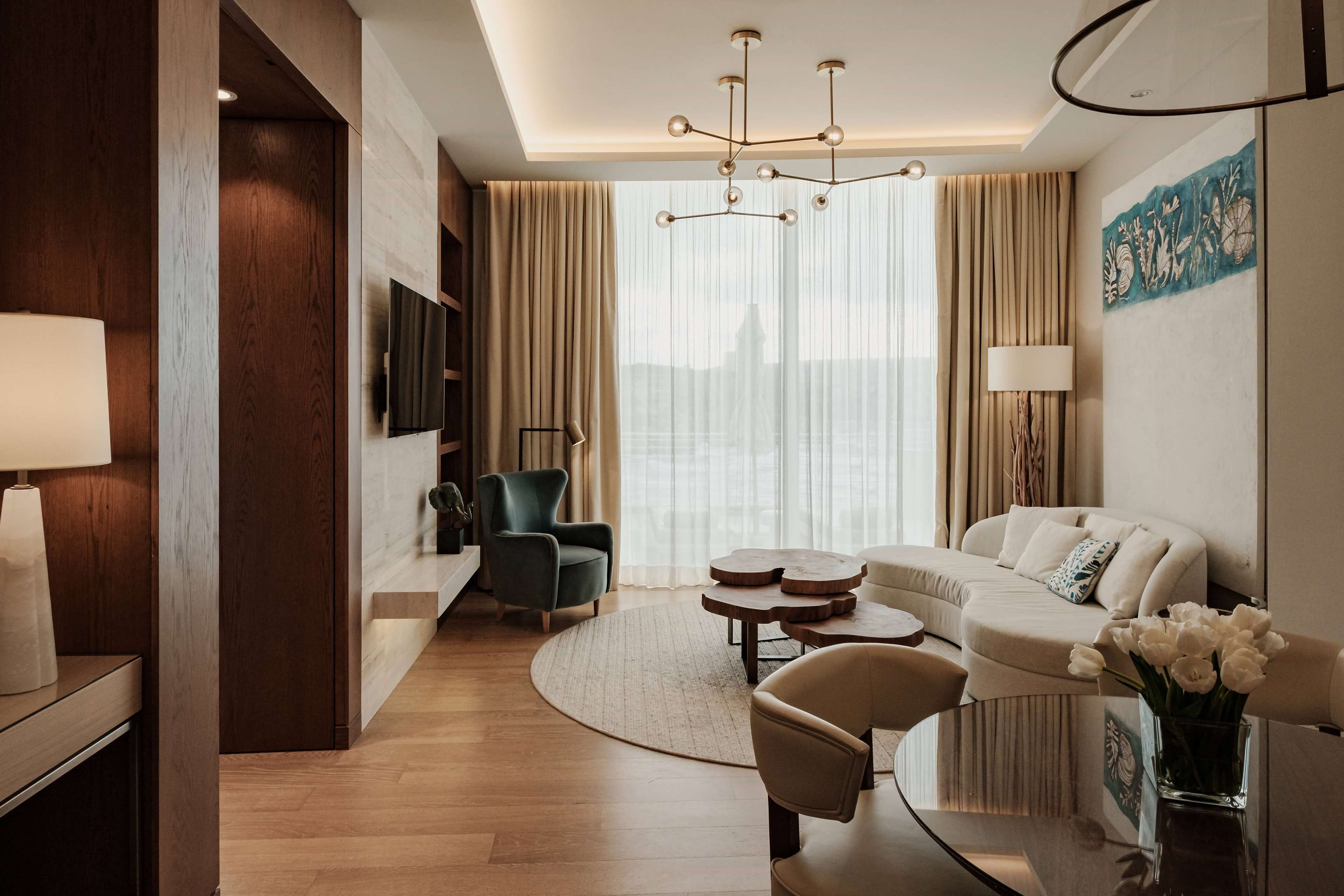 Una habitación de hotel moderna con una pared de acento de madera, un ventanal con cortinas translúcidas, una mesa de centro redonda, un sofá y un televisor montado en la pared.