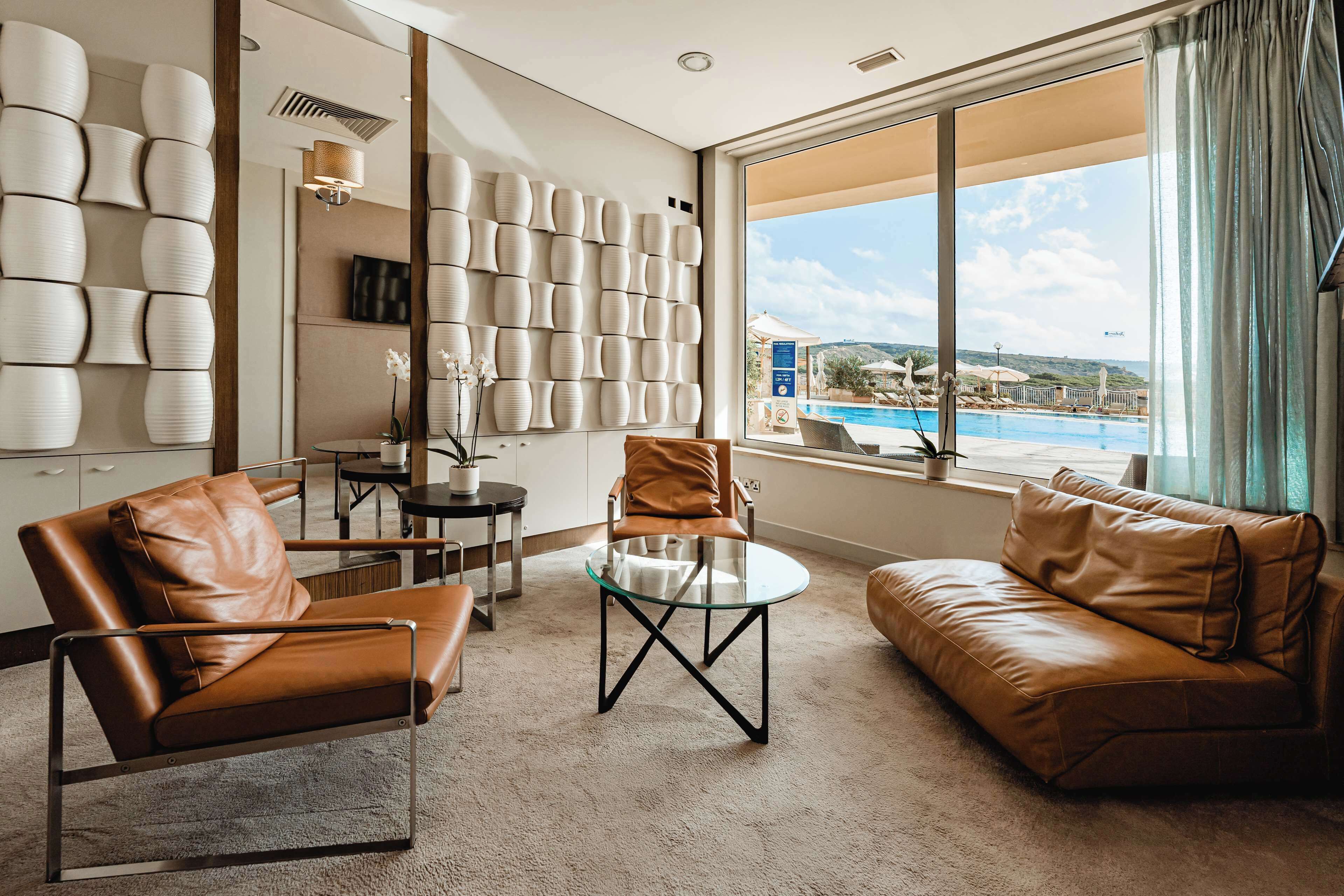 A modern hotel room with a leather sofa, a glass coffee table, and a view of a swimming pool.